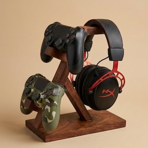 May include: A wooden gaming stand holds two black game controllers and a black and red gaming headset. The stand is made of dark brown wood and has a simple, modern design. The headset has the brand name on the side.