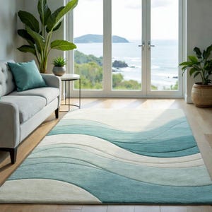 May include: A rectangular area rug with a wave design in shades of white, light blue, and teal. The rug is placed on a hardwood floor in a room with a sofa, plants, and a view of the ocean through large windows.