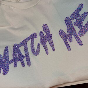 May include: A cream-colored t-shirt featuring the text WATCH ME spelled out in iridescent purple rhinestones. The letters are arranged in a slightly slanted, cursive-style font, with each individual stone reflecting light to create a shimmering effect.