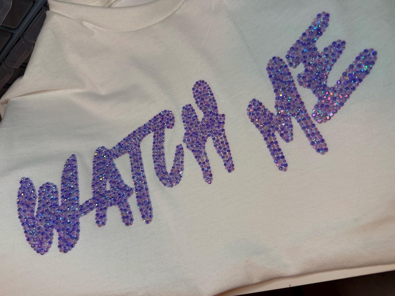 May include: A cream-colored t-shirt featuring the text WATCH ME spelled out in iridescent purple rhinestones. The letters are arranged in a slightly slanted, cursive-style font, with each individual stone reflecting light to create a shimmering effect.