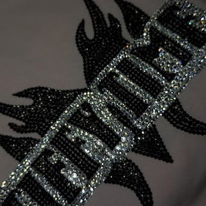 May include: A light-colored fabric garment featuring a decorative design composed of black and clear rhinestones. The design includes a flame-like shape in black rhinestones behind a rectangular border filled with clear rhinestones and the text HEAVEN.