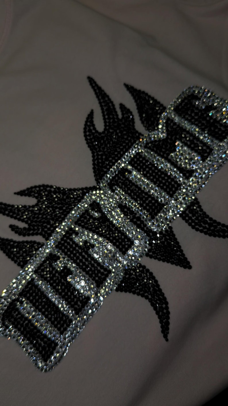 May include: A light-colored fabric garment featuring a decorative design composed of black and clear rhinestones. The design includes a flame-like shape in black rhinestones behind a rectangular border filled with clear rhinestones and the text HEAVEN.