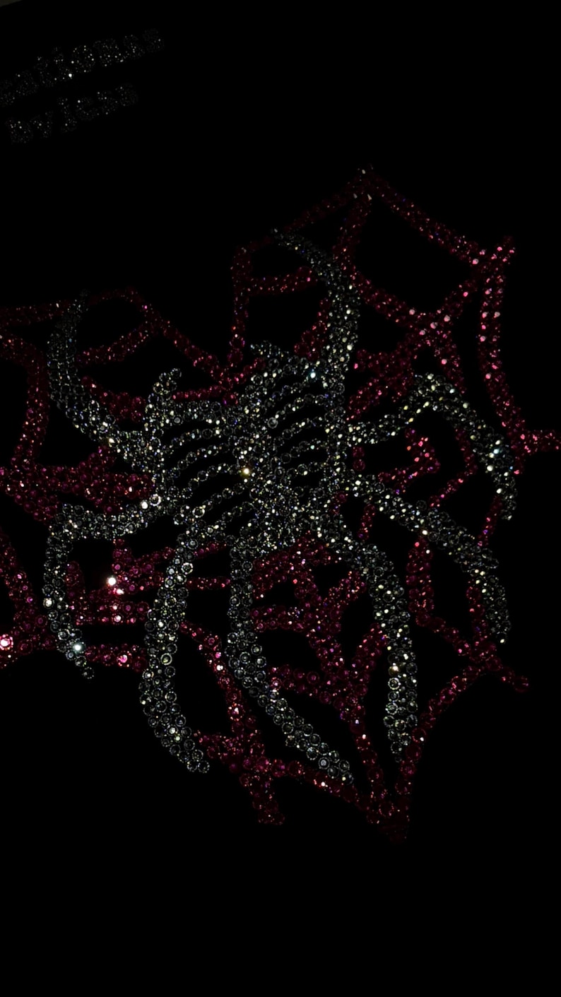 May include: A black fabric surface featuring a central spider design crafted from shimmering silver rhinestones, layered over a larger web pattern composed of deep red rhinestones. The design is set against a dark background with subtle text.