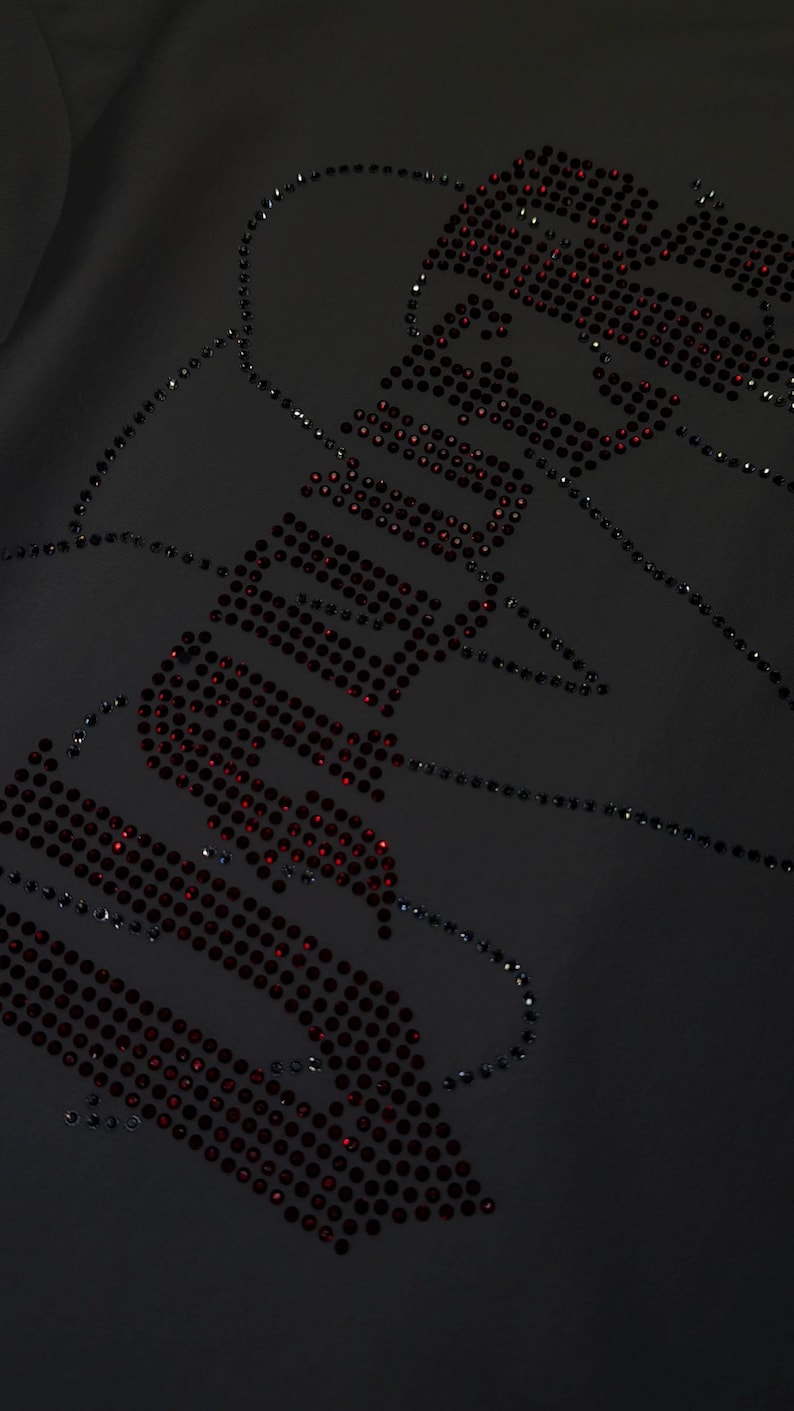 May include: A black fabric garment featuring a decorative design composed of small, dark red rhinestones. The rhinestones are arranged to form the word "SADBOYS" in a stylized, bold font, with additional scattered rhinestone accents.