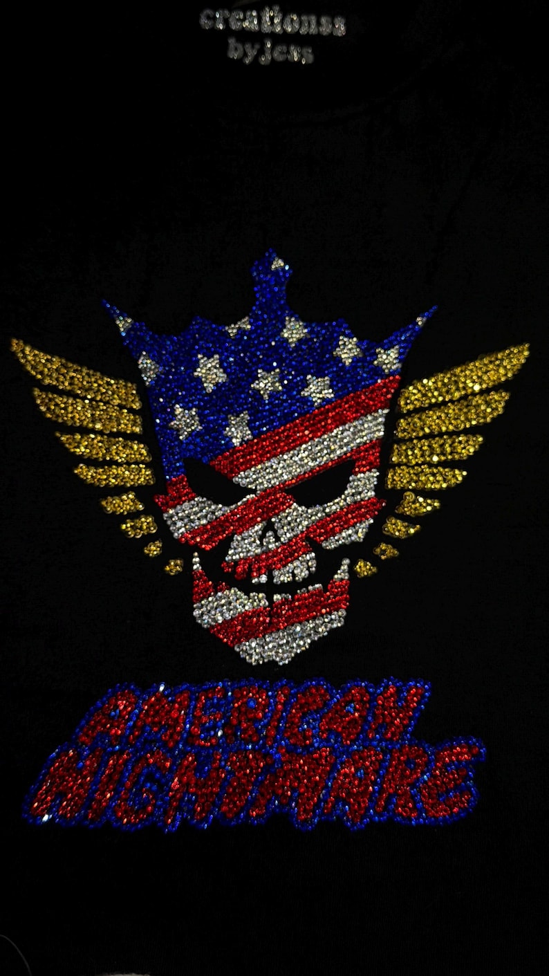 May include: A black garment featuring a rhinestone design of a skull wearing a crown, decorated with the American flag pattern and gold wings. Below the skull, the text AMERICAN NIGHTMARE is spelled out in red and blue rhinestones.