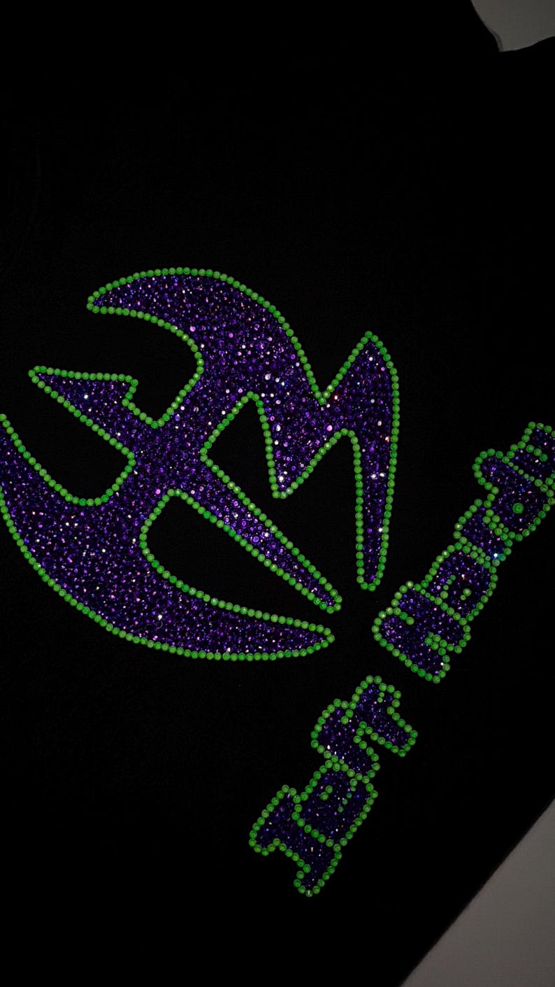 May include: A black garment featuring a large, stylized logo and the text Jeff Hardy. The design is created with purple rhinestones filled in the center and outlined with bright lime green rhinestones, creating a high-contrast, sparkling effect.