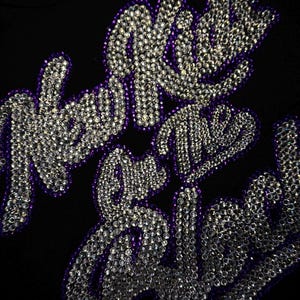 May include: A black fabric garment featuring the text New Kids on the Block spelled out in clear, reflective rhinestones. The lettering is outlined in a deep purple border, creating a high-contrast design against the dark background material.