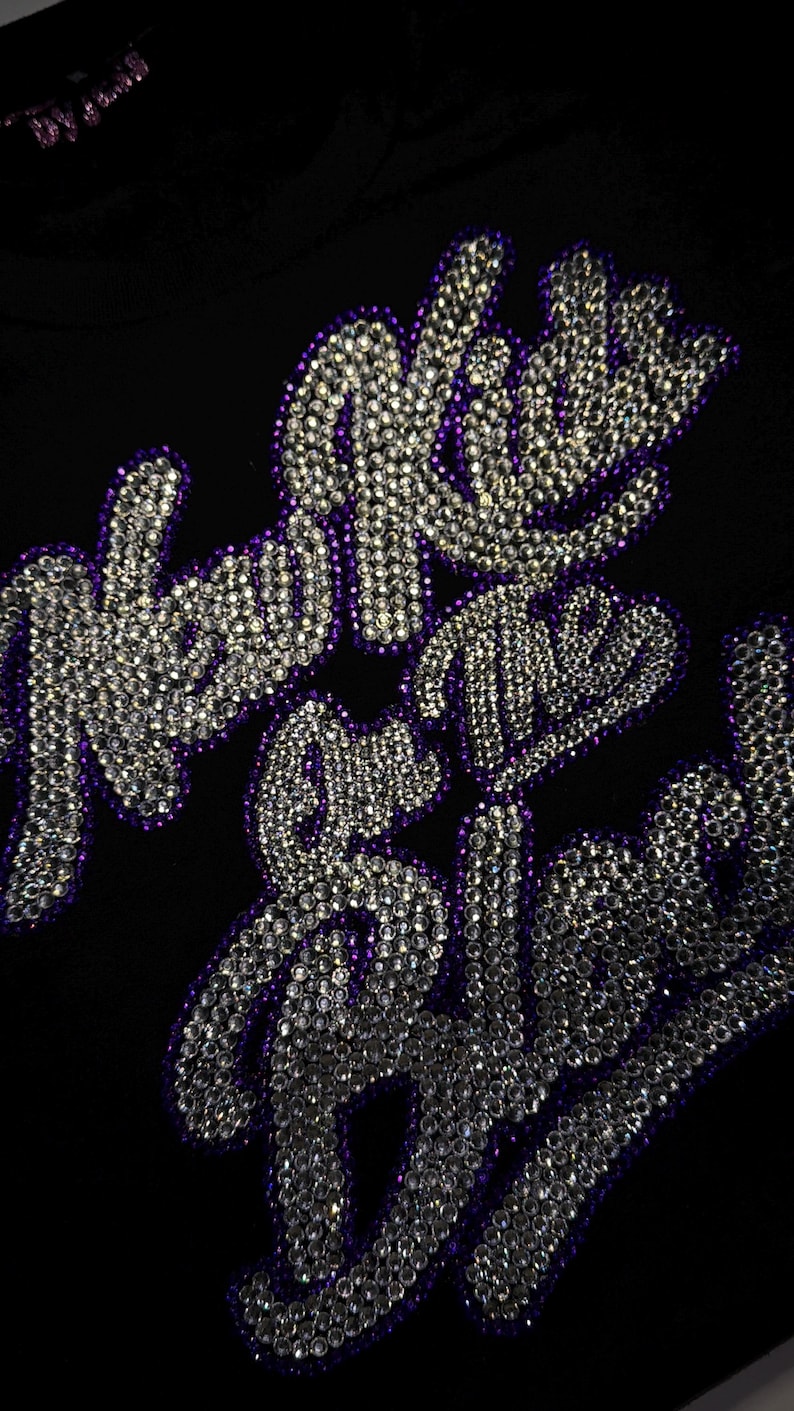 May include: A black fabric garment featuring the text New Kids on the Block spelled out in clear, reflective rhinestones. The lettering is outlined in a deep purple border, creating a high-contrast design against the dark background material.