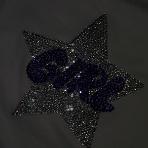 May include: A white t-shirt featuring a large star-shaped graphic made of clear, reflective rhinestones. The word "GIRL" is spelled out across the center of the star in dark purple rhinestones. The brand name "NO BOUNDARIES" is printed on the inner neck.