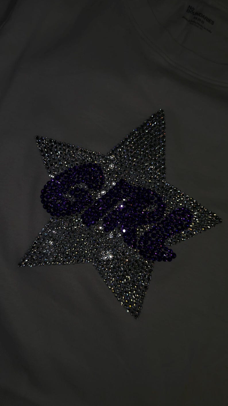 May include: A white t-shirt featuring a large star-shaped graphic made of clear, reflective rhinestones. The word "GIRL" is spelled out across the center of the star in dark purple rhinestones. The brand name "NO BOUNDARIES" is printed on the inner neck.