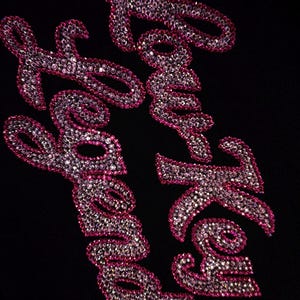 May include: A black fabric surface featuring the text "Low Key Legend" written in a cursive script. The letters are filled with small, reflective, clear crystals and outlined with a border of vibrant pink crystals, creating a sparkling effect.