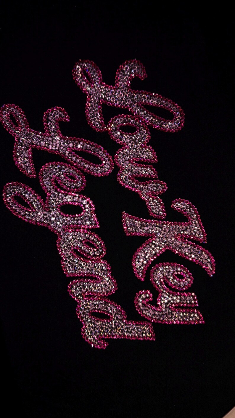 May include: A black fabric surface featuring the text "Low Key Legend" written in a cursive script. The letters are filled with small, reflective, clear crystals and outlined with a border of vibrant pink crystals, creating a sparkling effect.