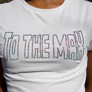 May include: A white short-sleeved t-shirt featuring the text TO THE MAX spelled out in iridescent, multi-colored rhinestones across the chest. The lettering is outlined in a bold, block-style font, creating a shimmering effect against the fabric.