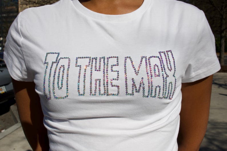 May include: A white short-sleeved t-shirt featuring the text TO THE MAX spelled out in iridescent, multi-colored rhinestones across the chest. The lettering is outlined in a bold, block-style font, creating a shimmering effect against the fabric.