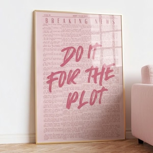 May include: A framed pink print with the words "BREAKING NEWS" at the top and "DO IT FOR THE PLOT" in a bold, pink script. The background resembles a vintage newspaper. The frame is gold, and a pink sofa is partially visible.