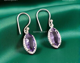 May include: Silver drop earrings featuring marquise-cut, purple gemstones. Each gemstone is framed by a halo of small, clear stones. The earrings are suspended from curved silver hooks, displayed on a dark green backdrop.