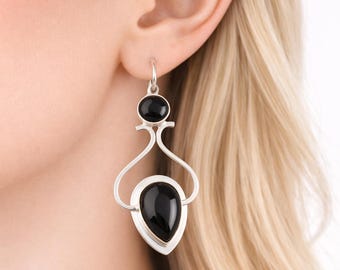 May include: A close-up of a silver earring with a teardrop-shaped black stone and a smaller, round black stone. The earring has a unique, open design with curved lines and a hook closure. The background is blurred, focusing on the jewelry.