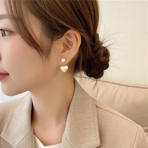 May include: A pair of pearl earrings. Each earring has a small pearl stud and a larger, heart-shaped pearl drop. The earrings are worn on a person's ear, with the focus on the jewelry. The background is blurred, highlighting the earrings.