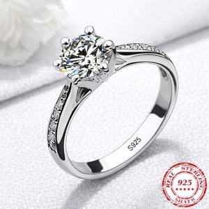 May include: A silver ring featuring a large, clear, round-cut gemstone. The band is embellished with smaller, clear gemstones. The ring is stamped with "S925" and a "Real Sterling 925 Silver" seal.