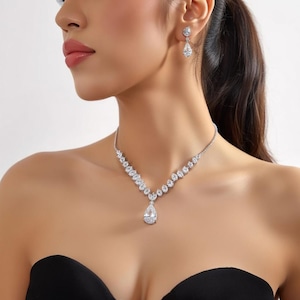 Puede incluir: Un conjunto de collar y pendientes plateados. El collar presenta una serie de pequeñas piedras redondas y transparentes que conducen a un gran colgante en forma de pera. Los pendientes tienen un diseño similar.