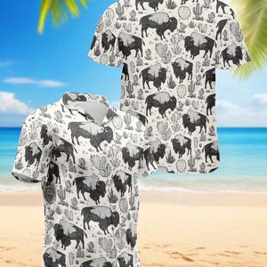 May include: Two short-sleeved button-down shirts with a repeating pattern of black and white bison and cacti on an off-white background. The shirts have a classic collar and are displayed against a beach backdrop.