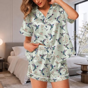 May include: A light green satin pajama set featuring a short-sleeved button-down shirt and matching shorts. The fabric is decorated with a repeating pattern of colorful hummingbirds and small floral sprigs, finished with white piping trim.