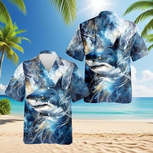 May include: Short-sleeved button-down shirt featuring a shark design. The shirt has a blue and white watercolor-style pattern with a shark graphic on the front and back. The background shows a beach scene with palm trees and the ocean.
