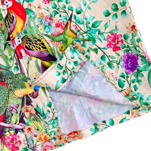 Peut inclure: Gros plan sur un tissu &agrave; imprim&eacute; tropical vibrant. Le motif pr&eacute;sente des perroquets color&eacute;s, un feuillage vert luxuriant et des fleurs aux couleurs vives dans les tons de rouge, de rose et de violet sur un fond cr&egrave;me. Le tissu est pli&eacute;.