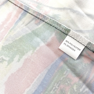 Peut inclure: Gros plan sur un tissu avec un motif abstrait color&eacute;. Le motif pr&eacute;sente des rayures m&eacute;lang&eacute;es de rose, de vert et de bleu. Une &eacute;tiquette blanche affiche "95% POLYESTER 5% SPANDEX". Le tissu est pli&eacute;, r&eacute;v&eacute;lant une couture noire.