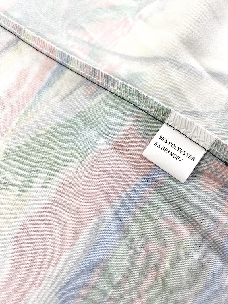 Peut inclure: Gros plan sur un tissu avec un motif abstrait color&eacute;. Le motif pr&eacute;sente des rayures m&eacute;lang&eacute;es de rose, de vert et de bleu. Une &eacute;tiquette blanche affiche "95% POLYESTER 5% SPANDEX". Le tissu est pli&eacute;, r&eacute;v&eacute;lant une couture noire.