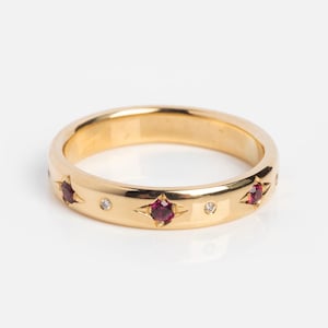 Op de afbeelding: Een gouden ring met stervormige robijnen en kleine diamantaccenten. De ring heeft een gepolijste afwerking en een klassiek bandontwerp. De edelstenen zijn gelijkmatig verdeeld.