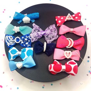 May include: A collection of colorful hair bows in various designs, including polka dots, lace, and glitter. The bows are in shades of blue, pink, and purple, with pearl and seashell accents. They are arranged on a black circular surface.