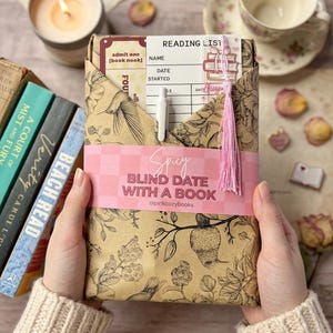 Könnte beinhalten: Ein eingepacktes Buch mit dem Text "Spicy Blind Date with a Book", das von Händen gehalten wird. Die Verpackung ist braun mit floralen und Vogelillustrationen. Ein rosa Band mit Text umwickelt das Buch. Eine Leseliste und ein Stift sind sichtbar.