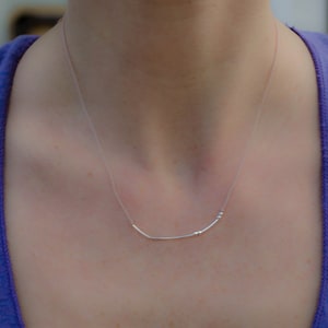 May include: A delicate silver necklace with a curved bar pendant and two small beads. The necklace is worn against a person's skin, with a purple tank top visible in the background. The necklace is simple and elegant.