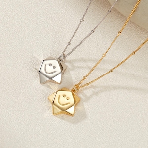 May include: Two necklaces with star-shaped pendants, one silver and one gold. Each pendant features a smiling face design. The necklaces have delicate chains with small bead accents. The background is a light, textured surface.