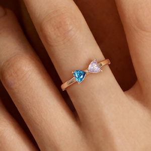 May include: A rose gold ring featuring two heart-shaped gemstones, one blue and one pink. The ring is displayed on a person's finger, showcasing the delicate design and color contrast of the gemstones. The ring is a piece of jewelry.