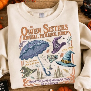 May include: A cream-colored crewneck sweatshirt featuring a graphic print with the text "Owen Sisters Annual Parasol Jump" and "Rooftop Jump & Margaritas at Midnight". The design includes a purple parasol, witch hats, candles, and a margarita glass.