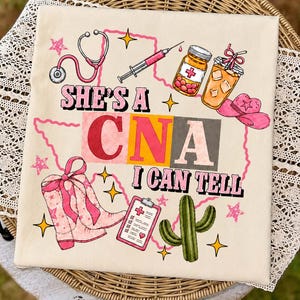 Può includere: Una t-shirt color crema con una stampa grafica che riporta il testo 'SHE'S A CNA I CAN TELL'. Il design include uno stetoscopio rosa, una siringa, un flacone di pillole, una bevanda, un cappello da cowboy rosa, stivali rosa, una cartella clinica e un cactus.