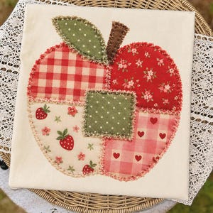 May include: A square off-white fabric pillow cover featuring a large apple-shaped applique made of various patterned fabric patches. The apple design includes red gingham, floral prints, strawberry motifs, and polka dots with frayed edges.