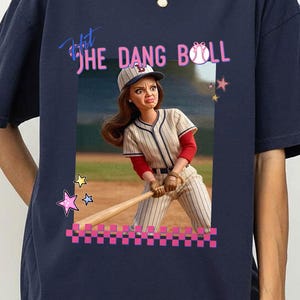 May include: Navy blue t-shirt featuring a graphic of a baseball player with the text "Hit The Dang Ball". The graphic includes a baseball, stars, and a pink checkered border. The baseball player is wearing a baseball uniform.