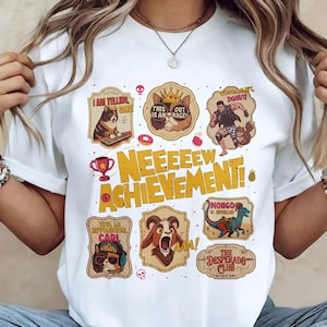 May include: White t-shirt featuring a variety of cartoon animal graphics with text. The shirt has the words "NEEEEEW ACHIEVEMENT!" in large yellow letters. Other graphics include a cat, a dog, a dinosaur, and a ram.