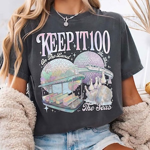 May include: Dark gray t-shirt with the text "KEEP IT 100" above a graphic of a theme park with a boat ride, a geodesic sphere, and the text "On The Land" and "The Seas". The shirt has a vintage, faded look.