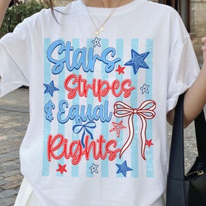 以下が含まれることがあります： 白のTシャツ。赤と青で「Stars, Stripes & Equal Rights」の文字と星、リボンが描かれた愛国的なデザイン。薄い青のストライプの背景。