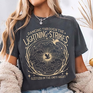 Könnte beinhalten: Dunkelgraues T-Shirt mit dem Text "Dancing Through The Lightning Strikes" und "Sleepless in the Onyx Night". Das Design zeigt eine kreisförmige Grafik mit einem Vogel, Wolken und Blitzen in Gold.