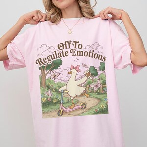 Pode incluir: Camiseta rosa claro com a frase "Off To Regulate Emotions". O gráfico apresenta um pato de desenho animado andando de patinete rosa, segurando uma xícara de café e usando um laço. O design inclui borboletas e um caminho.