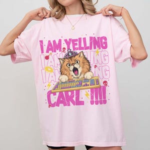 May include: Pink t-shirt with a graphic of a cat wearing a crown and typing on a keyboard. The text on the shirt reads "I AM YELLING CARL!!!" in pink and white. The cat has brown and tan fur and a surprised expression.