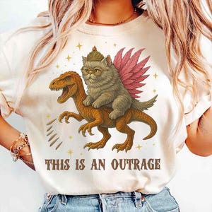 May include: Cream-colored t-shirt featuring a whimsical illustration. A cat wearing a crown and glasses with pink wings rides a dinosaur. The text "THIS IS AN OUTRAGE" is printed below the image. Gold stars are scattered around the design.