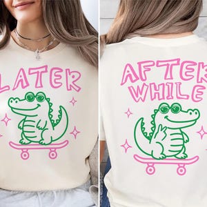 Peut inclure: T-shirt crème avec un alligator vert portant des lunettes de soleil sur un skateboard rose. Le devant du t-shirt dit "LATER" en rose, et le dos dit "AFTER WHILE". Le design comprend des étoiles roses.