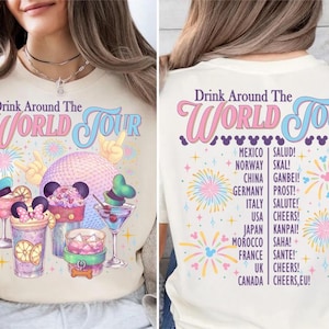 May include: Cream-colored t-shirt with the text "Drink Around The World Tour" in pink and blue. The front features colorful illustrations of drinks and a large sphere. The back lists countries and greetings.