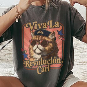 May include: A charcoal gray short-sleeve t-shirt featuring a graphic of a fluffy brown cat wearing sunglasses, a headset, and a blue cap with a crown. The text reads Viva La Revolucion, Carl, surrounded by red and blue stars on a peach background.